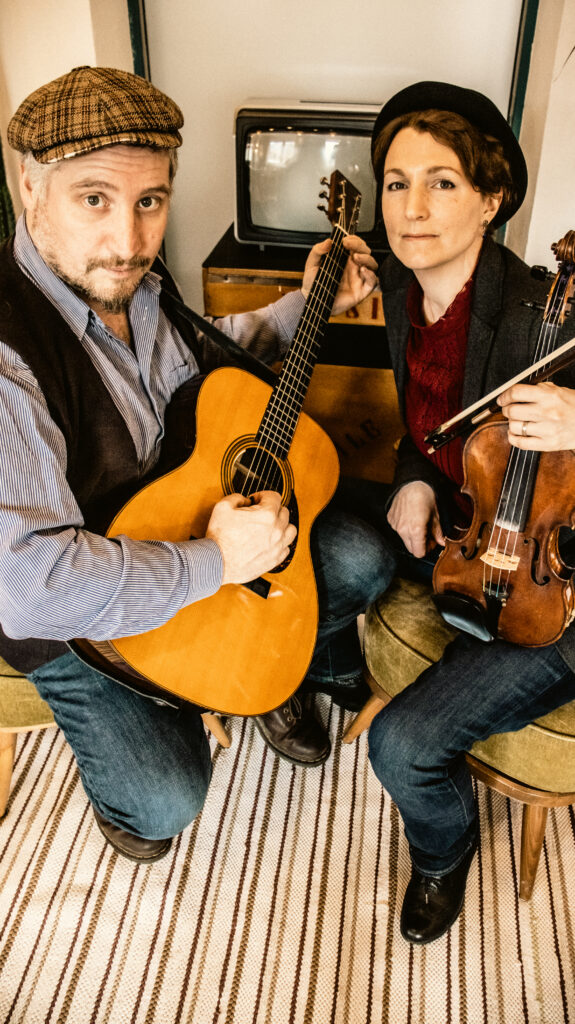 Dominik Plangger sitzt mit Gitarre auf dem Schoß neben Claudia Fenzl, die ihre Geige in der Hand hält.