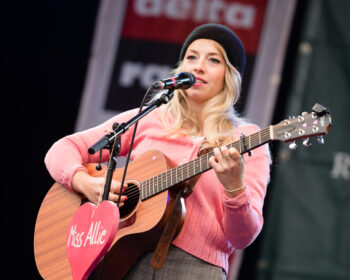 Miss Allie, eine blonde Frau mit pinkem Pullover und Beanie, steht mit einer Gitarre an einem Mikrofon an dem ein ebenfalls pinkes Herz hängt