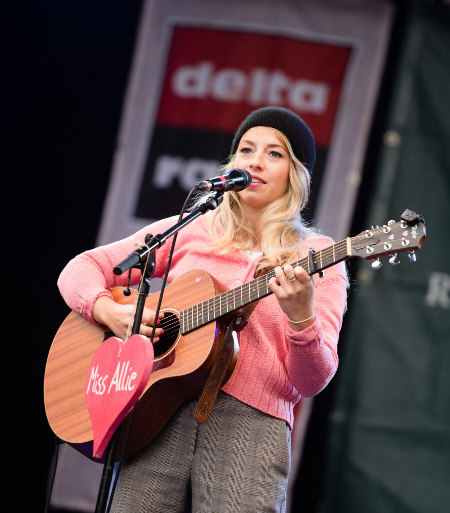 Miss Allie, eine blonde Frau mit pinkem Pullover und Beanie, steht mit einer Gitarre an einem Mikrofon an dem ein ebenfalls pinkes Herz hängt
