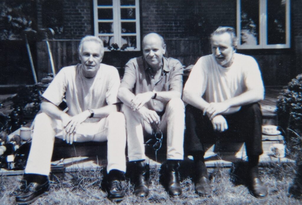 Ein schwarz-weiß Bild mit drei Männern, die vor einem Haus hocken und in die Kamera blicken